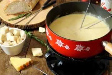 a red caquelon filled with real swiss cheese fondue on a wooden board surface surrounded by various ingredients