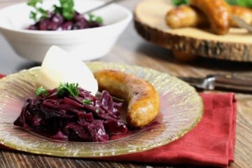 braised german red cabbage recipe served on a fancy gold rimmed glass plate alongside sausages and mashed potatoe.