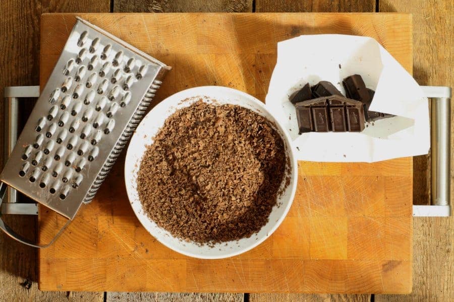 A box grater , white bowl, and dark bakers chocolate laying on a wooden cutting board. The bowl is filled with freshly grated chocolate shavings.