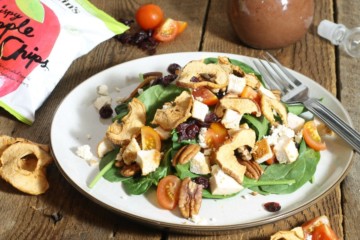 a chicken spinach salad garnished with crispy apple chips, feta, cherry tomatoes and pecans on a wooden table