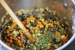 raw wild rice, herbs, and garlic being added to the pot of sauteed vegetables