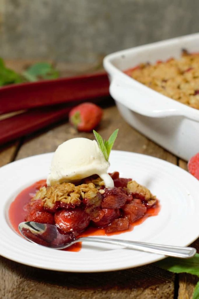 vertical image of strawberry rhubarb crumble served with vanilla ice cream and mint  on a white plate