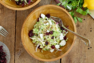 Overhead shot of shaved brussels sprout salad in a rustic wooden bowl, garnished with dried cranberries and goat cheese