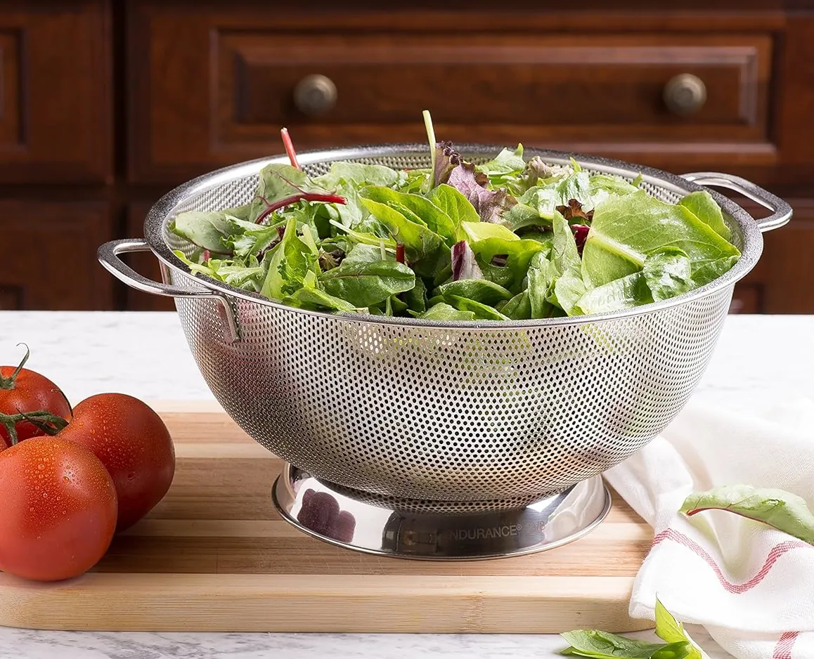 Stainless steel colander with handles full of green leaves standing on the wooden board with tomatoes