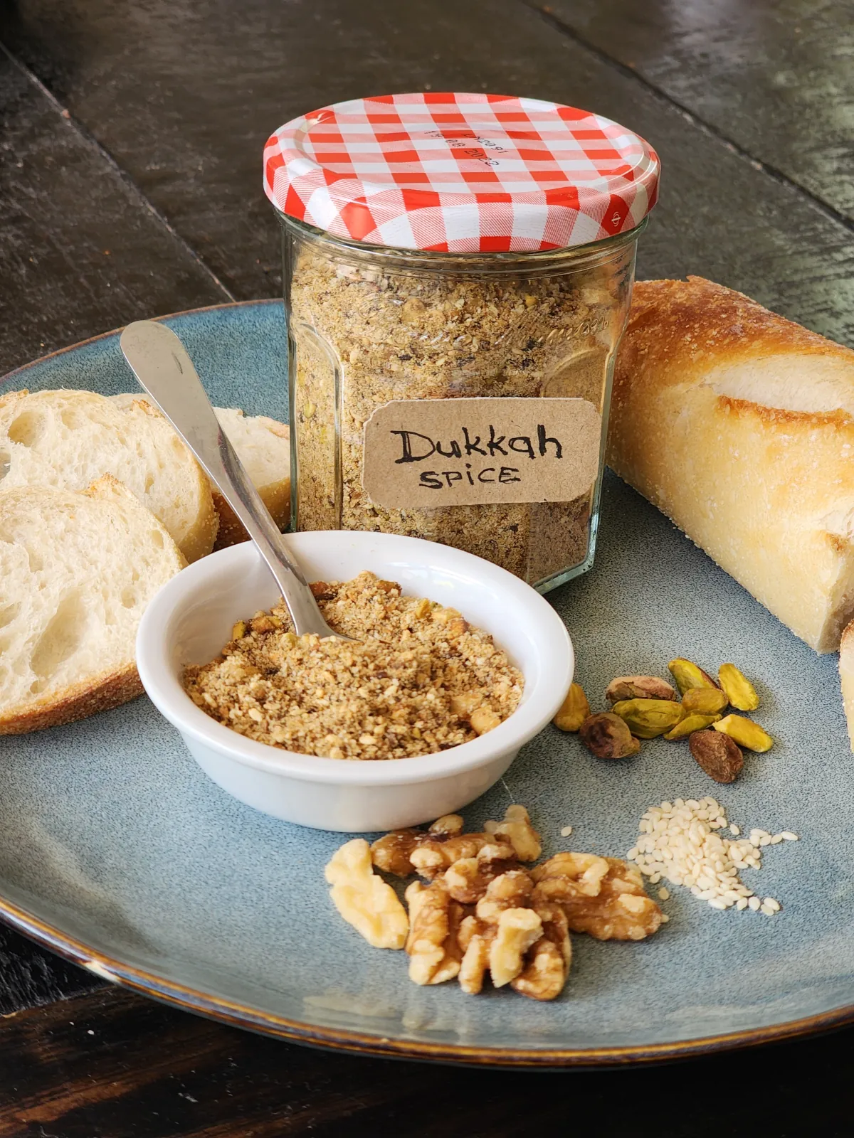 Homemade Dukkah Spice in a jar and a small dish on dinner plate with baquette, walnut and pistachio nuts and sesame seeds.